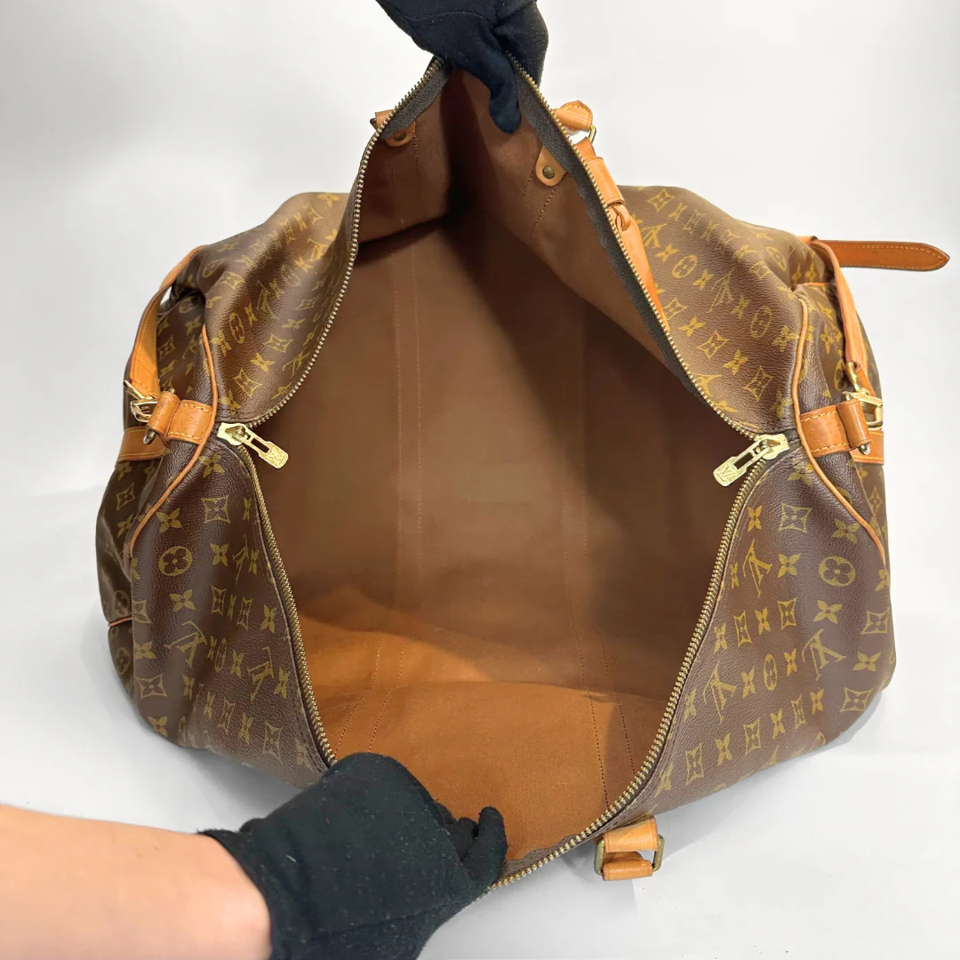 Brown monogrammed handbag with gold accents held by a gloved hand on a white background